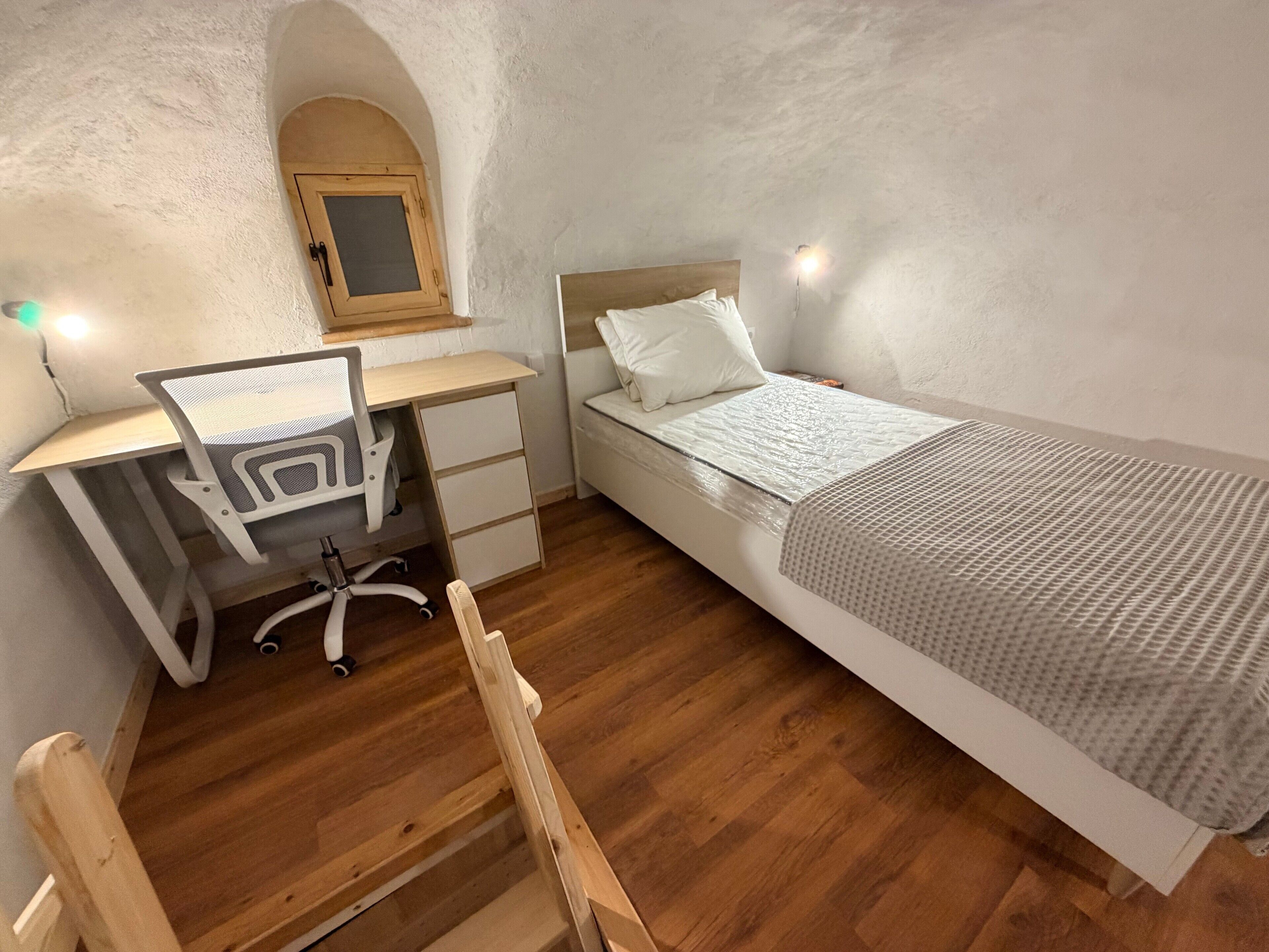 Une chambre avec un bureau en bois, un lit, une chaise et une petite fenêtre en arc de cercle.
