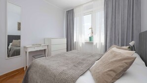 Une chambre à coucher avec un lit, un bureau, une chaise, un miroir et une fenêtre avec des rideaux.