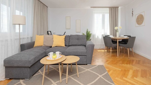 Un salon moderne avec un canapé d’angle gris, une table basse ronde en bois et un coin repas comprenant une petite table et des chaises.