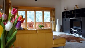 Una sala de estar con un sofá amarillo, una chimenea, un televisor y un ventanal con vistas a árboles.