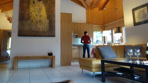 Un amplio salón con techo alto, vigas de madera, un cuadro grande, un sofá, una mesa de centro y una cocina al fondo.