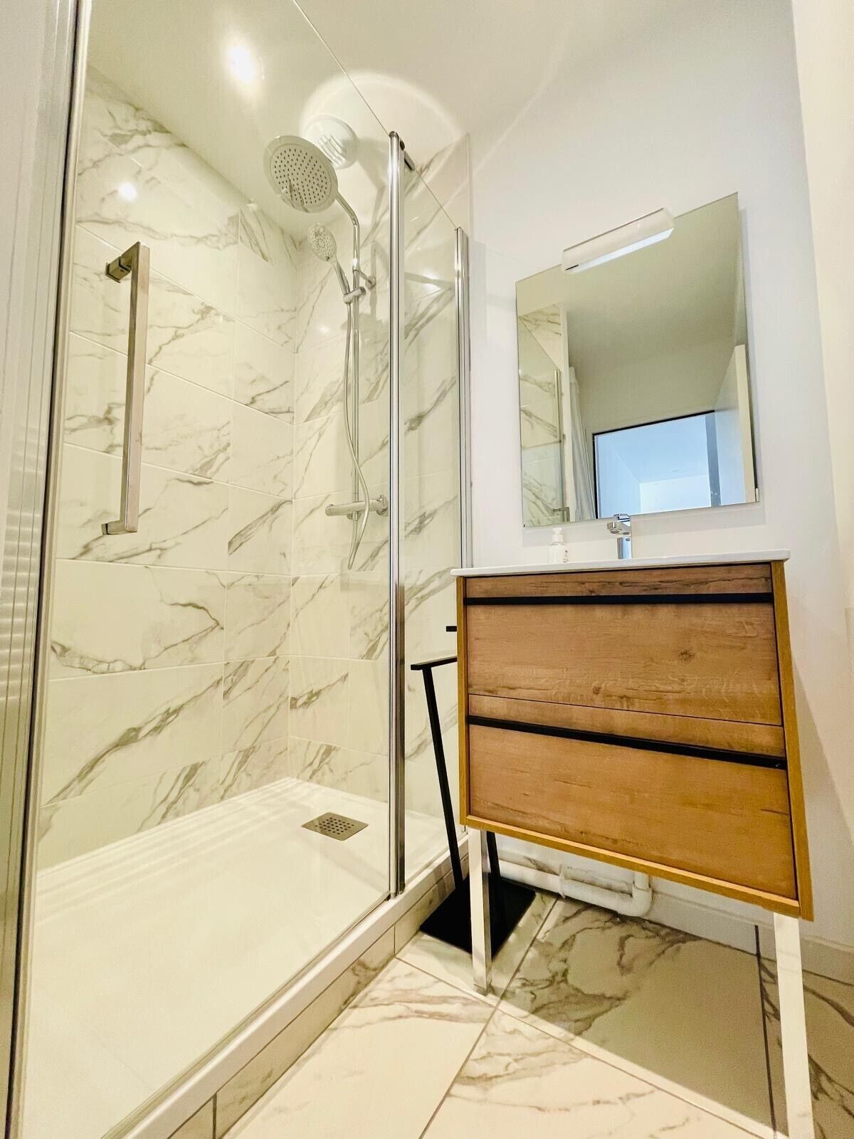 A modern bathroom with a glass-enclosed shower, marble tiles, a wooden vanity, and a mirror above the sink.