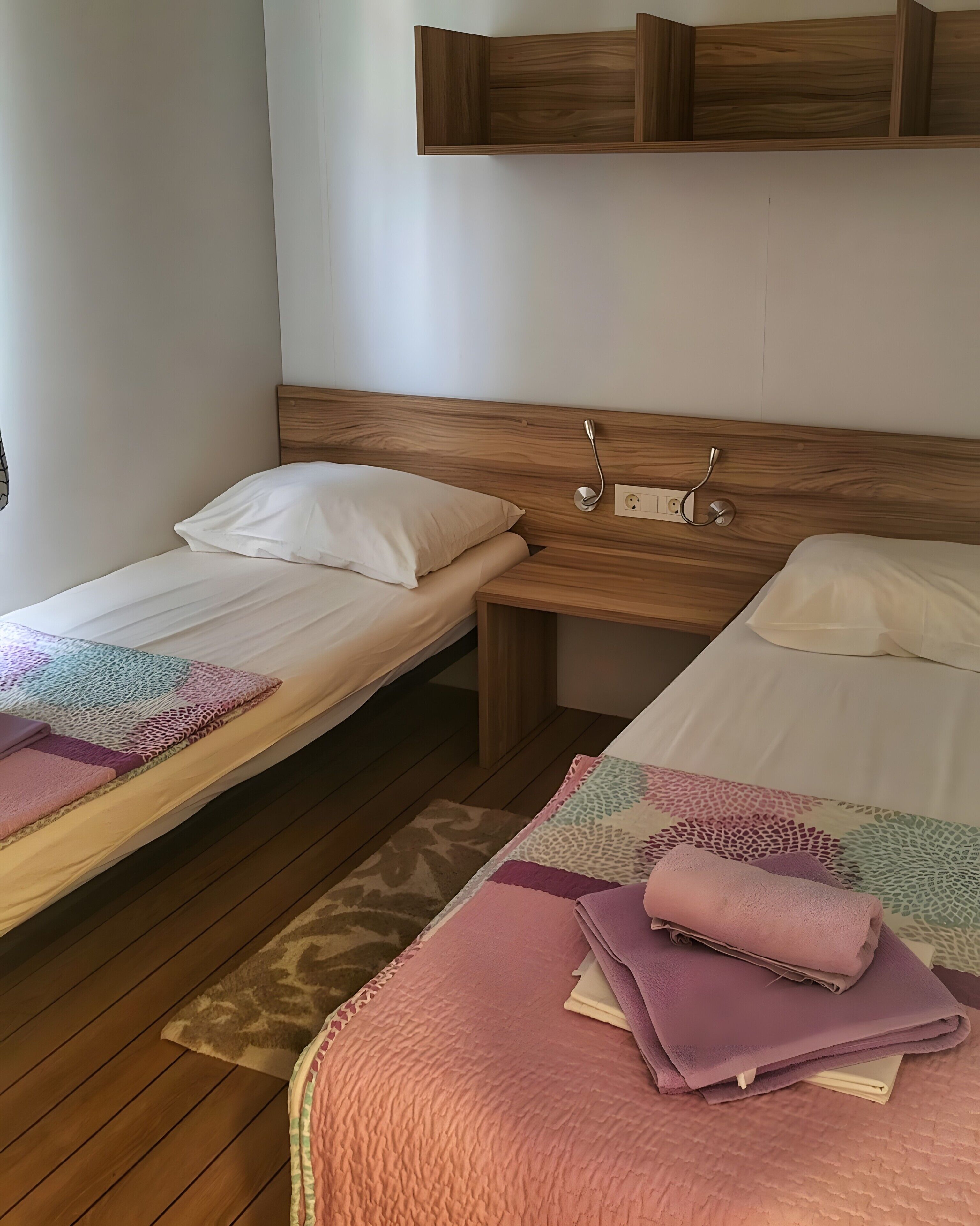 A room with two single beds, wooden headboards, a bedside table, and a wall-mounted shelf.