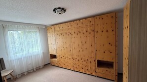 Ett rum med en stor garderob i trä, ett litet träbord och ett fönster med tunna, genomskinliga gardiner.