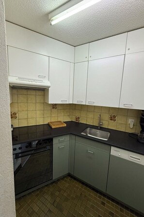 A compact kitchen with white cabinets, a black countertop, a stove, a sink, and a dishwasher. - Parsennresidenz 403 Appartement/Fewo, Dusche oder Bad, WC 2½-Zimmerwohnung by Interhome (Davos)