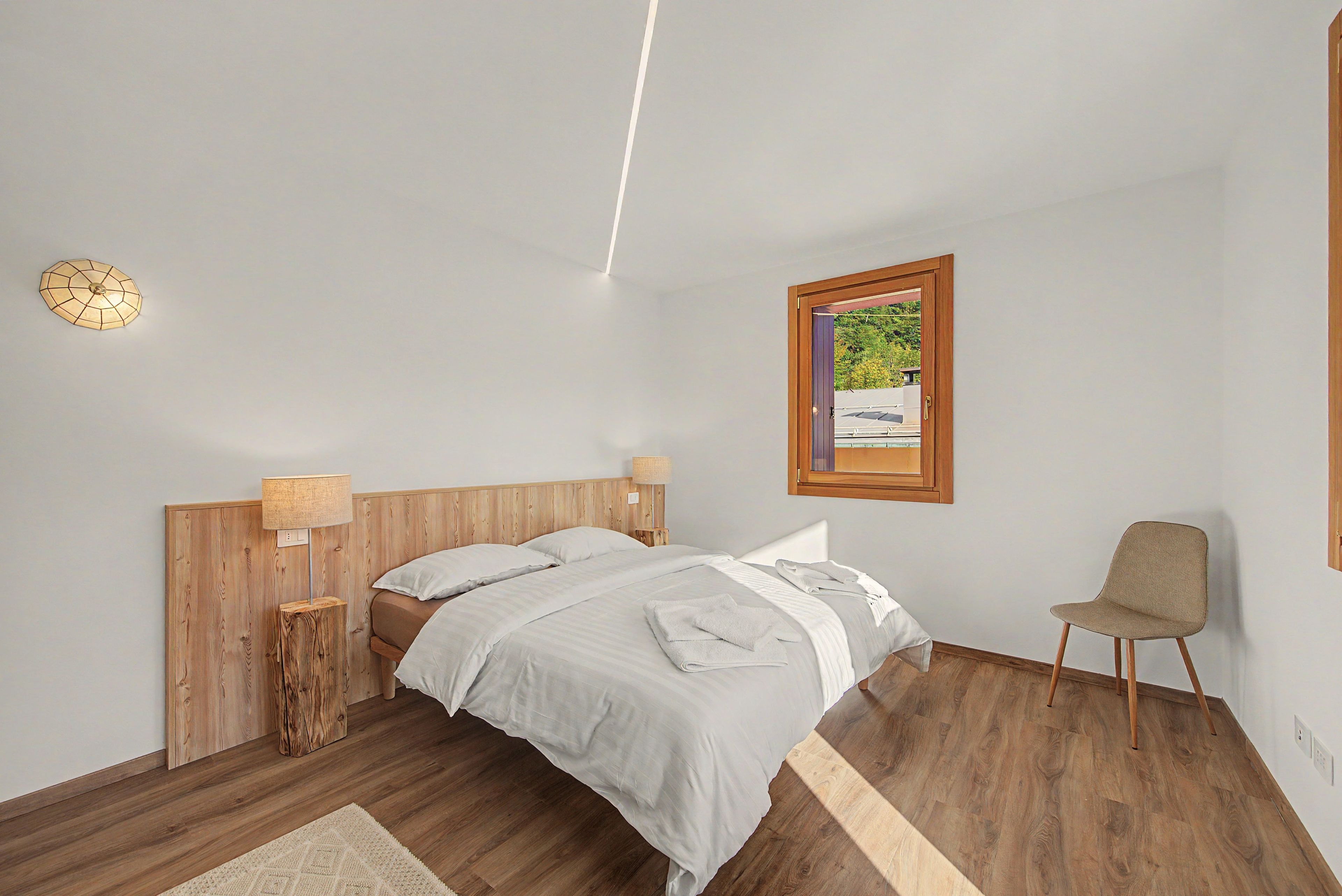 Ein Schlafzimmer mit einem hölzernen Kopfende, zwei Nachttischlampen, einem Bett mit weißen Leinen, einem Holzboden, einem kleinen Teppich und einem Sessel.