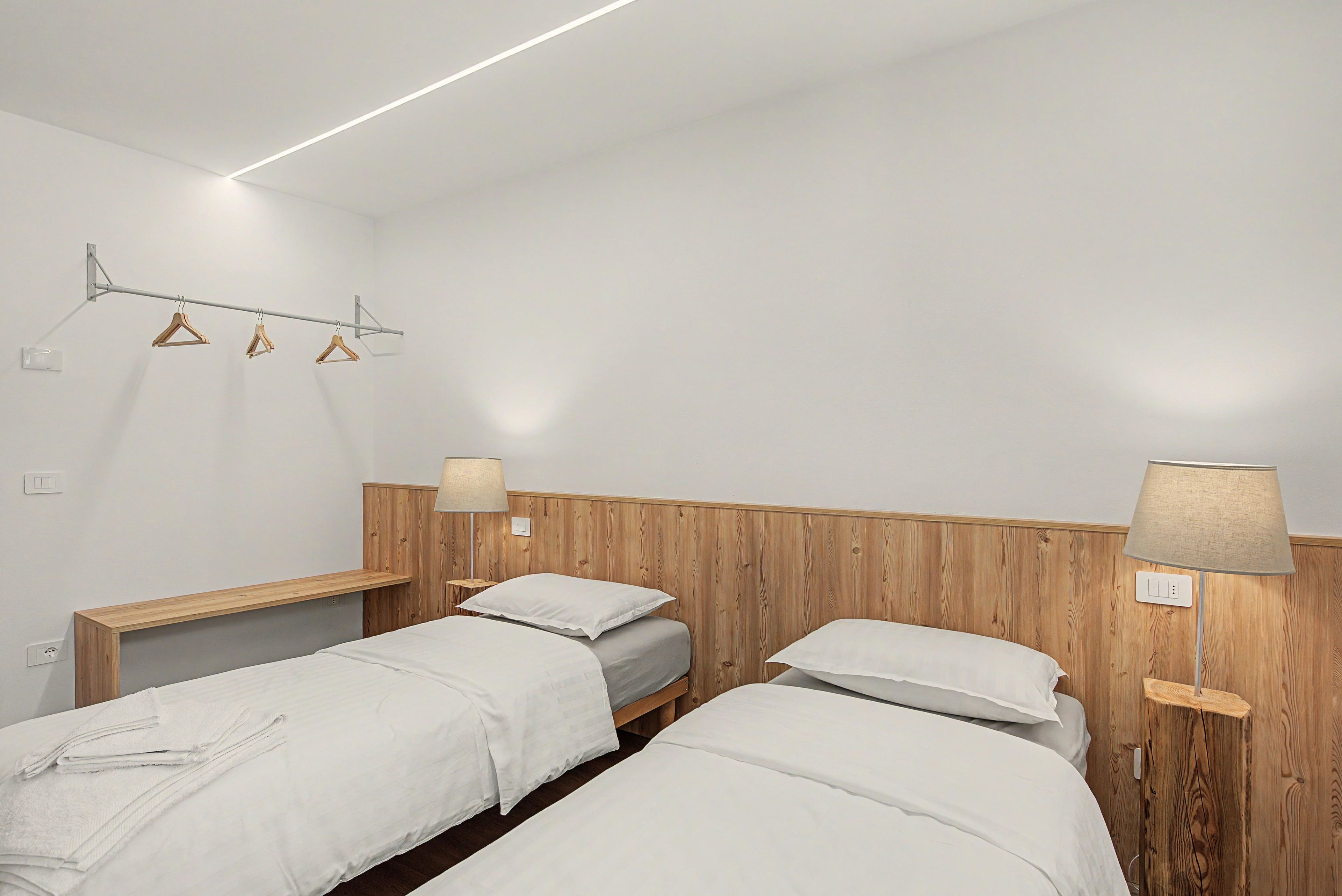 A hotel room with two beds, wooden headboards, bedside lamps, and a wall-mounted drying rack.