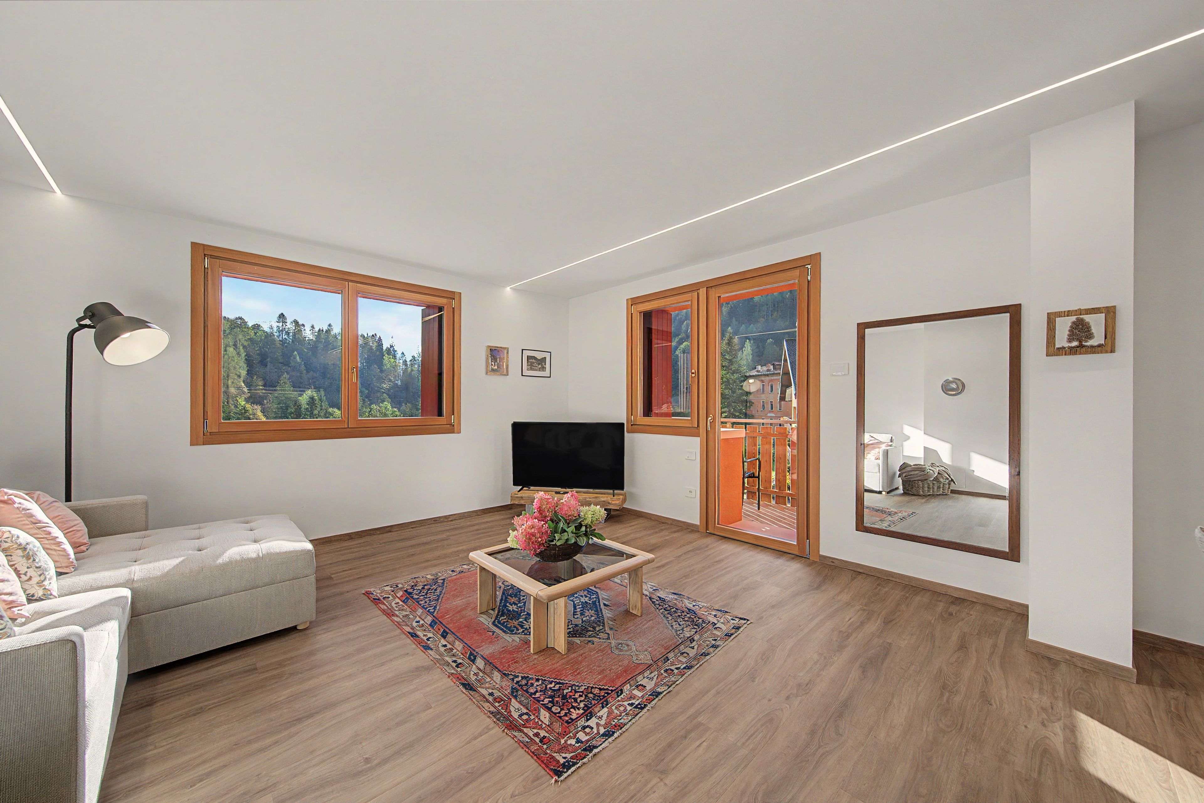 A modern living room with a sofa, a coffee table with flowers, a TV, and large windows with a view of trees.