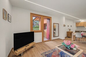A modern living room with a flat-screen TV on a wooden stand, a dining area with a table and chairs, and a kitchen area with wooden cabinets.