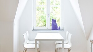 A room with a white table and chairs, a window with a view of greenery, and a purple object on the windowsill. - Stadt Land Fluss Studio Apartment '2' in Regensburg (Regensburg)