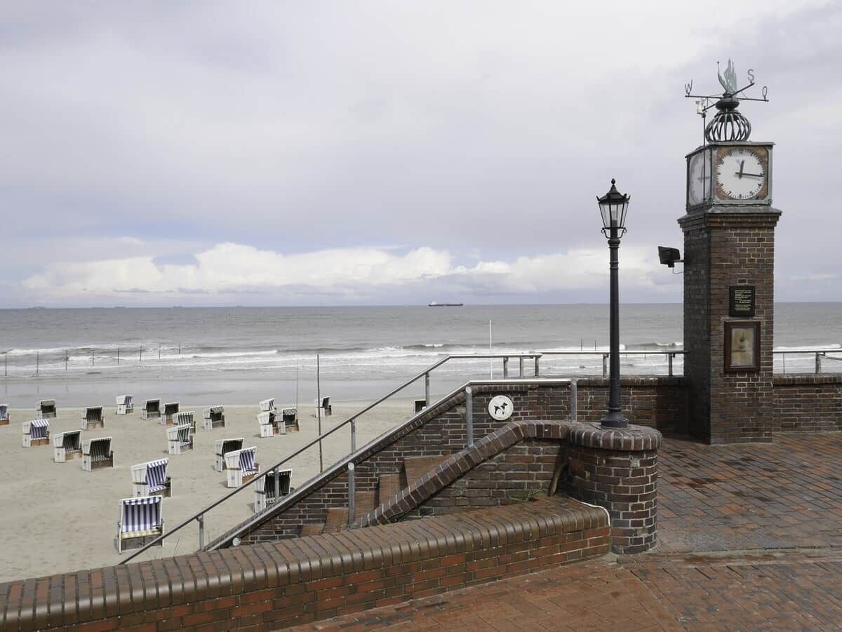 Eine Strandpromenade mit einem Glockenturm, Treppen zu Liegestühlen und Blick auf das Meer.