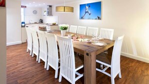 Uma área de refeições com uma mesa de madeira, cadeiras brancas e uma cozinha ao fundo.