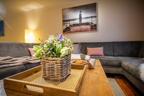 Ein Wohnzimmer mit einem Korbbusch aus Blumen auf einem Tablett, einem grauen Sofa, einem Holztisch und einem gerahmten Bild an der Wand.