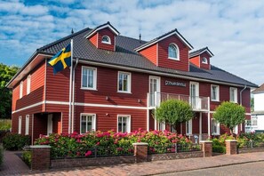 A red building with a flag, labeled 'Schwedenhus,' with a brick pathway and flower beds.