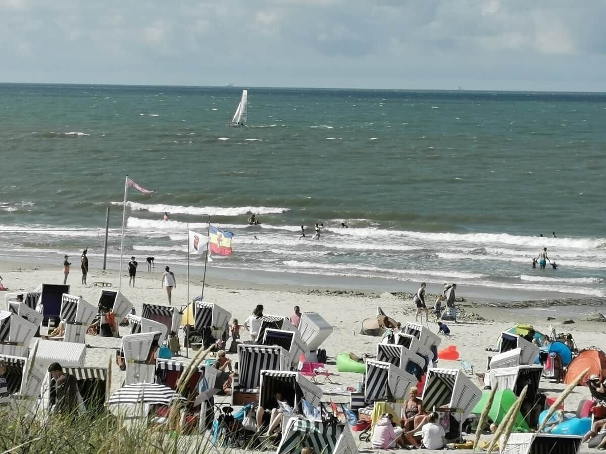 Een strand met rijen ligstoelen en parasols, een zeilboot op het water, en mensen in de verte.