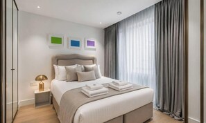 A modern bedroom with a bed, bedside tables, and a window with curtains. - Riverside Studio Manhattan Sleeps 2 - Chelsea AC (London)