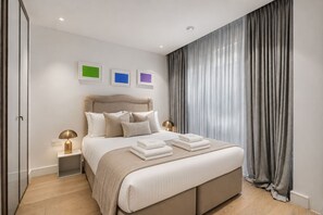 A modern bedroom with a large bed, bedside lamps, and a view of the outside through sheer curtains. - Riverside Studio Manhattan Sleeps 2 - Chelsea AC (London)
