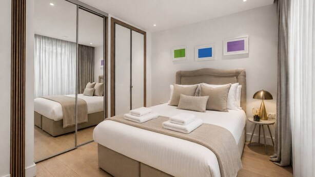 A modern hotel room with a large bed, a mirror, a bedside table with a lamp, and three framed pictures on the wall. - Riverside Studio Manhattan Sleeps 2 - Chelsea AC (London)