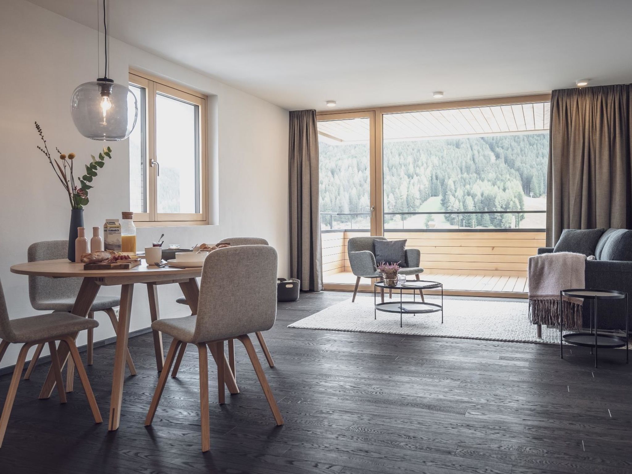 A modern living room with a dining area, large windows with a view of a forest, and a cozy seating area.