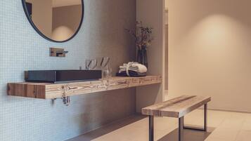 Une salle de bain moderne avec un meuble-lavabo suspendu, un miroir rond, un banc et un vase rempli de fleurs.