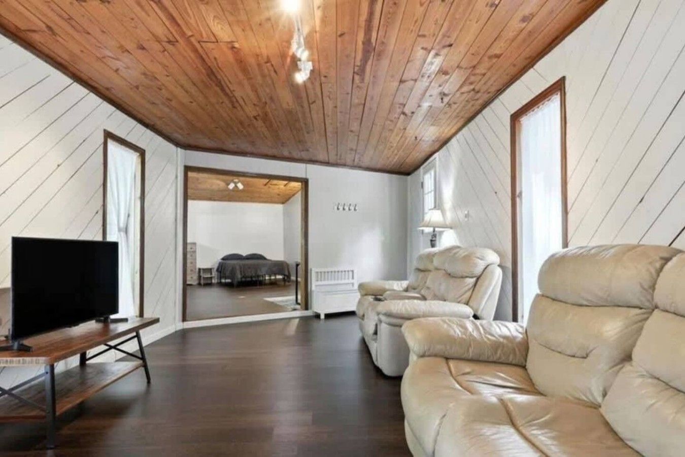 A room with a wooden ceiling, a leather sofa, a television, and a bed in the background.