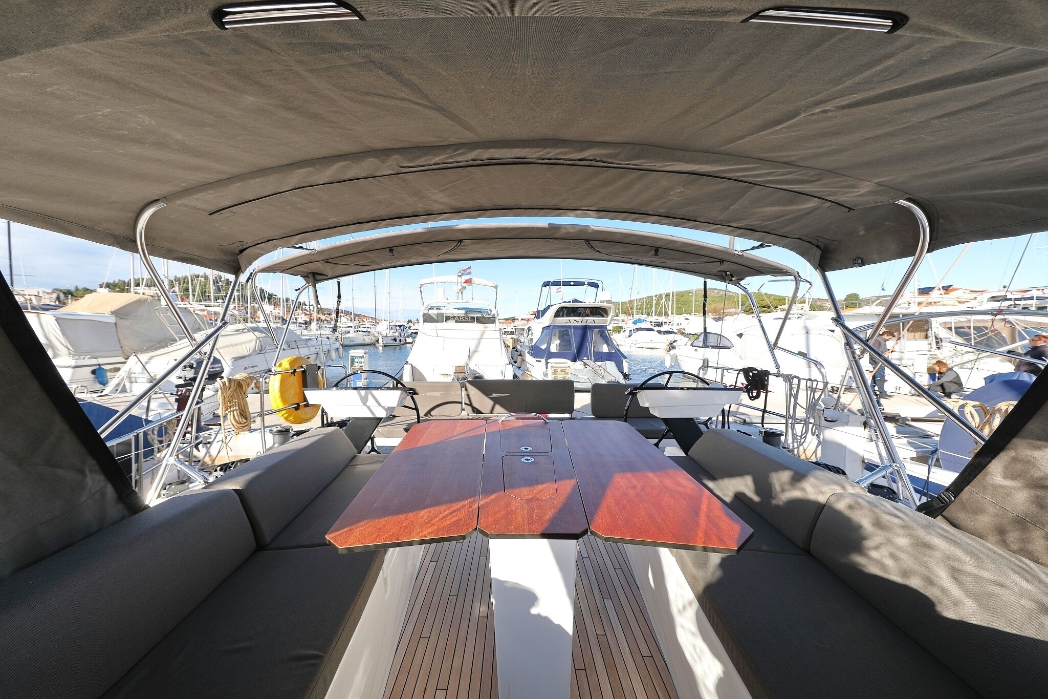 A boat's interior with a cushioned seating area, a table, and a view of other boats at a marina.