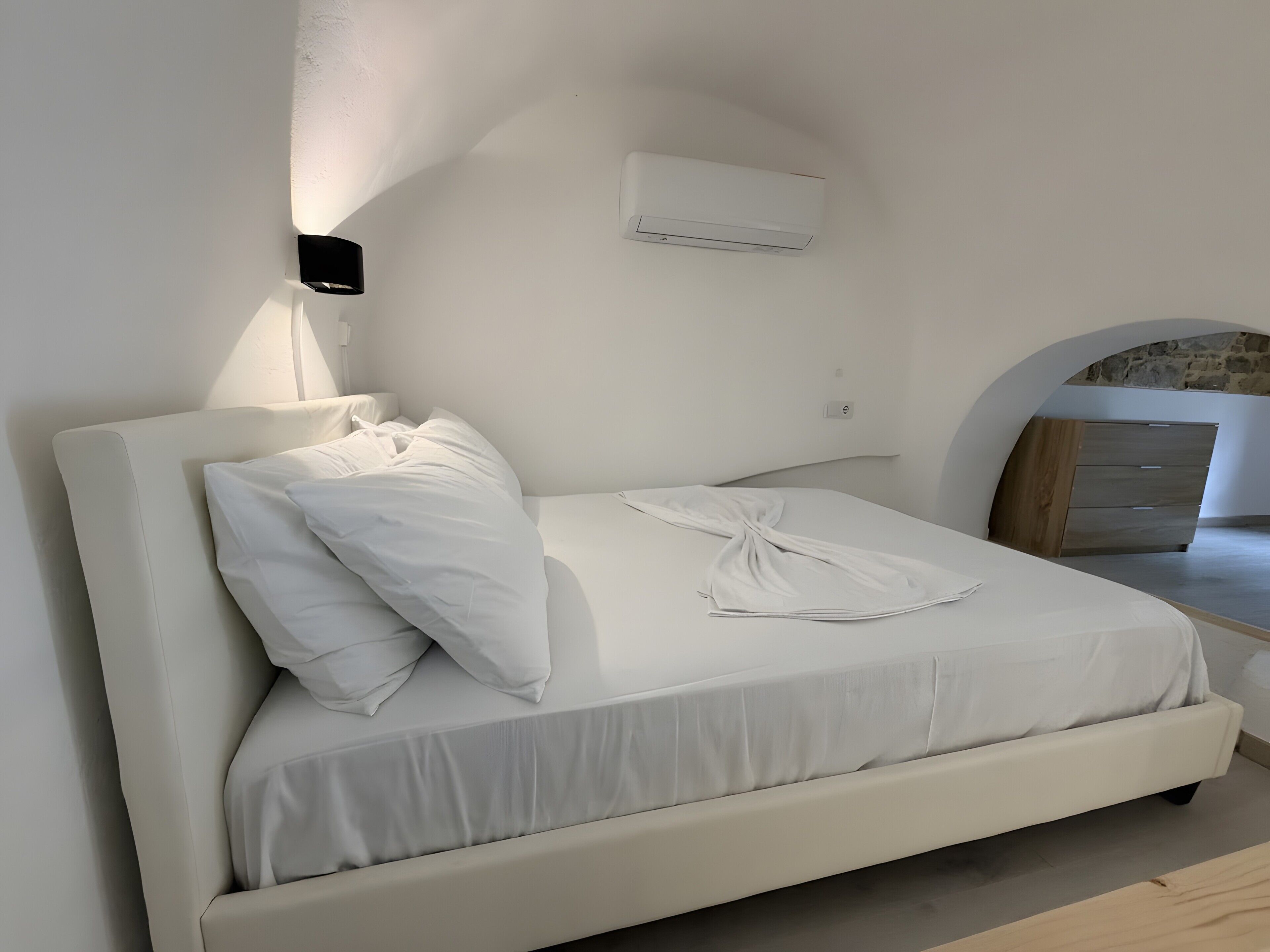 Un lit avec du linge de lit blanc, un climatiseur fixé au mur et une armoire en bois.
