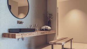 Un baño moderno con un mueble de tocador suspendido, un espejo redondo, una banca y un florero con flores.