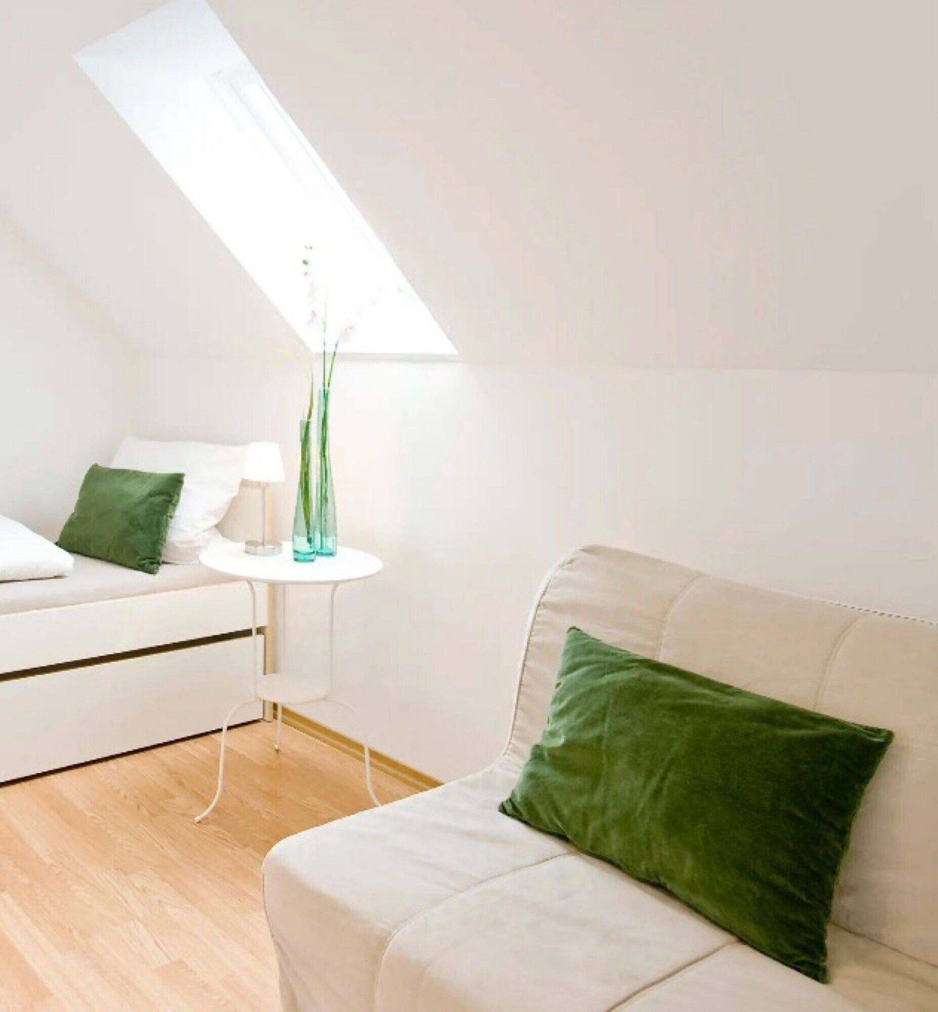 Una habitación con techo inclinado, un sofá blanco con un cojín verde, una mesita de noche blanca con un florero, y un aparador blanco.
