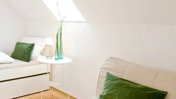 Una habitación con techo inclinado, un sofá blanco con un cojín verde, una mesita de noche blanca con un florero, y un aparador blanco.