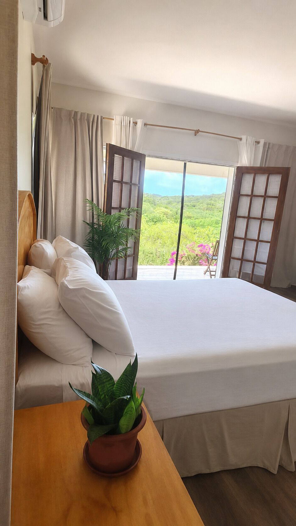 A bedroom with a large bed, a wooden nightstand, a potted plant, and a view of a green landscape through a window.