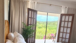 Una camera da letto con un letto grande, un comodino in legno, una pianta in vaso e vista su un paesaggio verde attraverso una finestra.