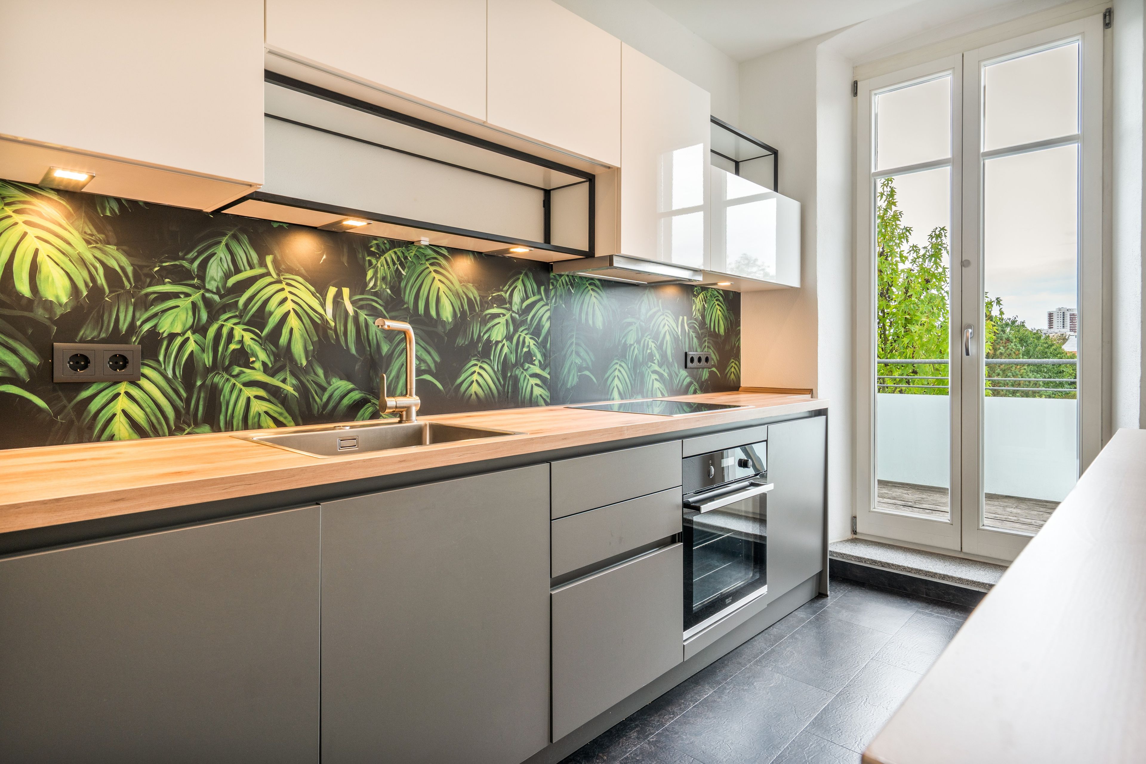 Una cocina moderna con un ventanal grande, horno empotrado y salpicadero con diseño de hojas tropicales.