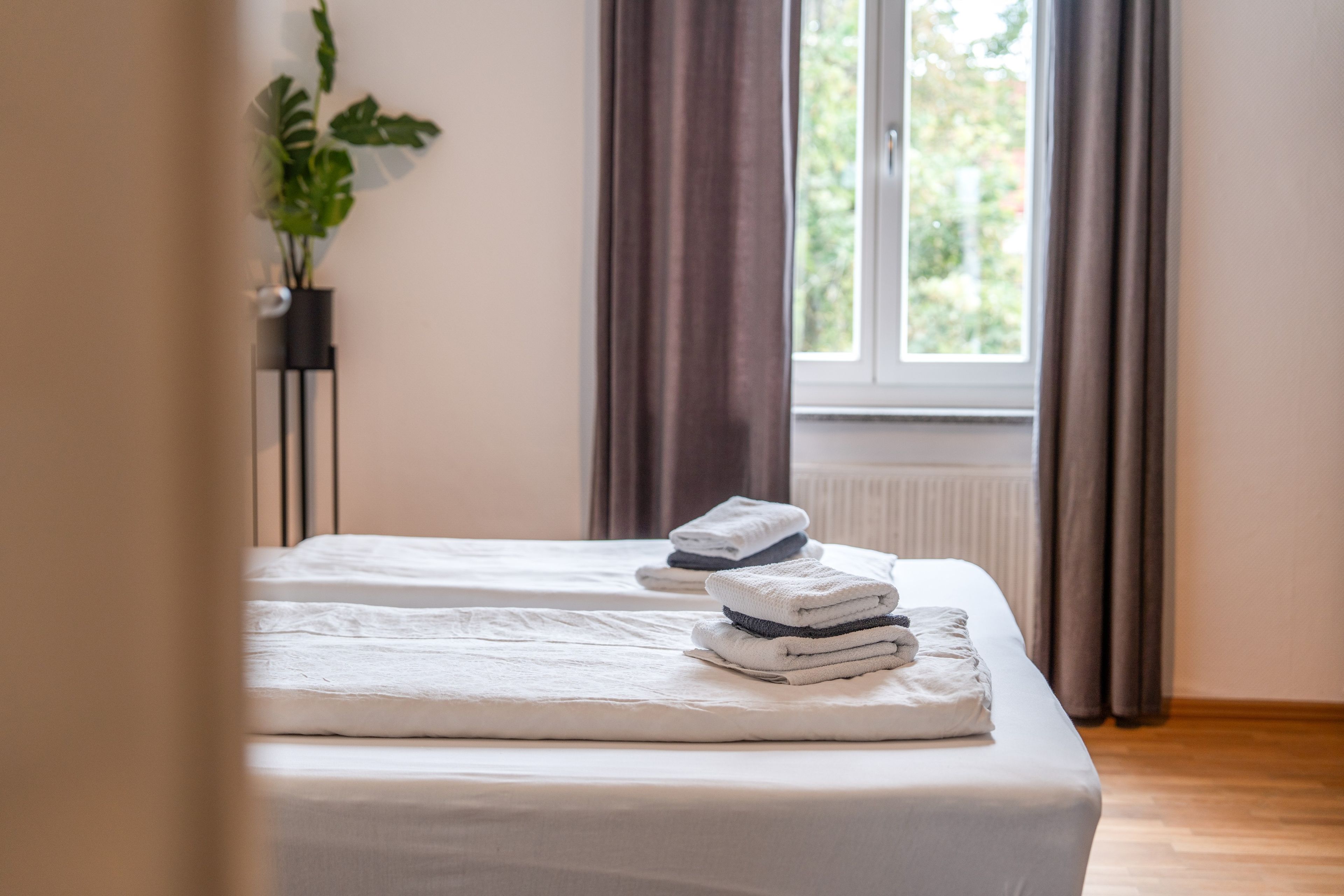 Ein ordentlich bezogenes Bett mit gefalteten Handtüchern, einer Topfpflanze und einem Fenster mit Vorhängen.