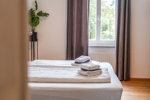 Ein ordentlich bezogenes Bett mit gefalteten Handtüchern, einer Topfpflanze und einem Fenster mit Vorhängen.