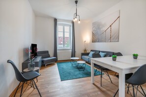 A modern living room with a sofa, a dining table, and chairs. There is a window with curtains, a painting on the wall, and a potted plant.