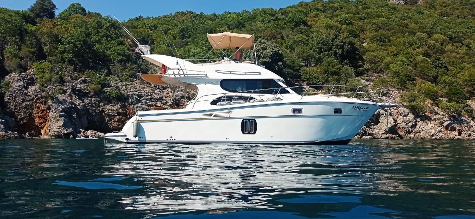 A white yacht with a canopy, anchored near rocky terrain and dense greenery.