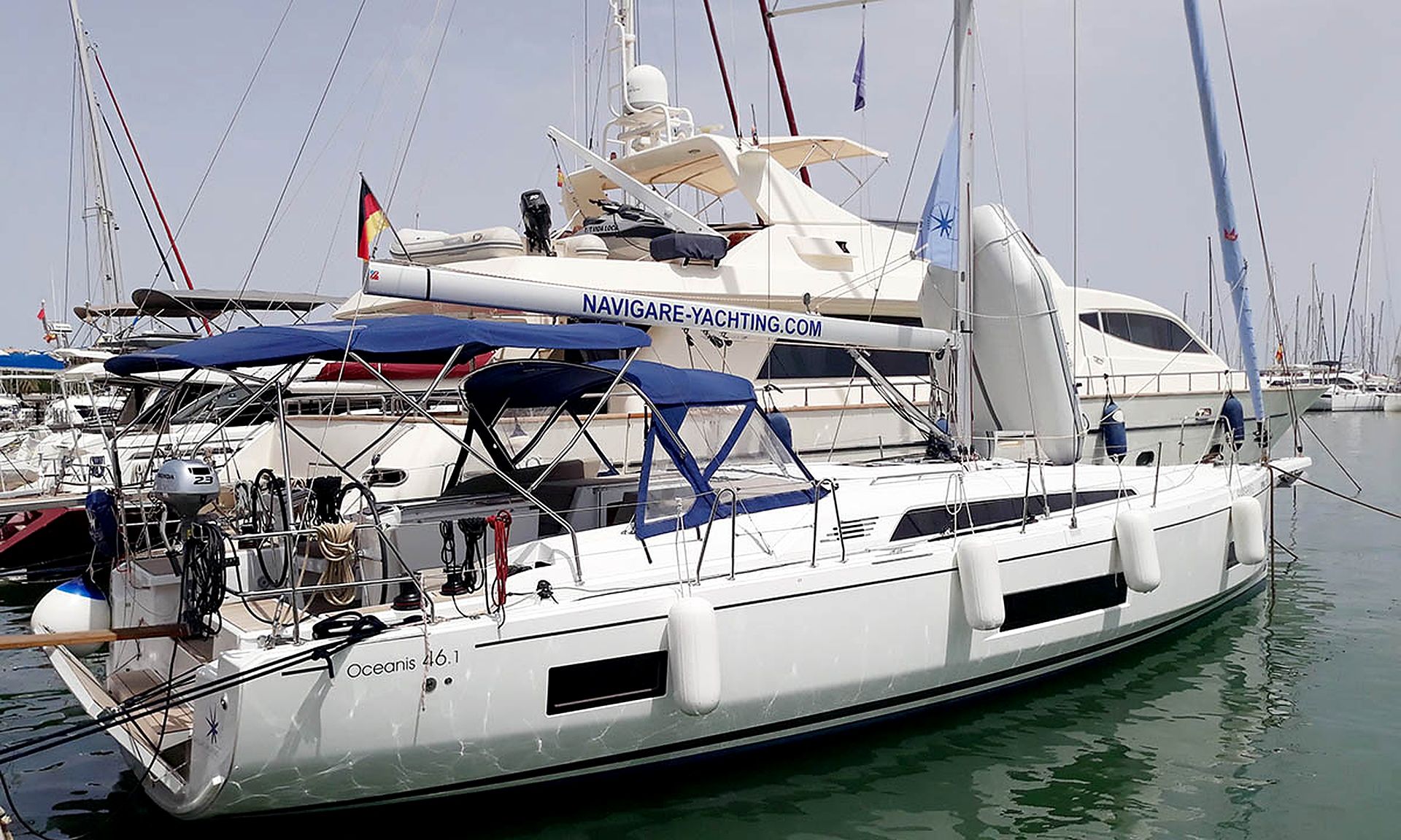 A yacht docked at a marina with multiple flags, a website banner, and the name 'Oceanis 46.1' on the hull.