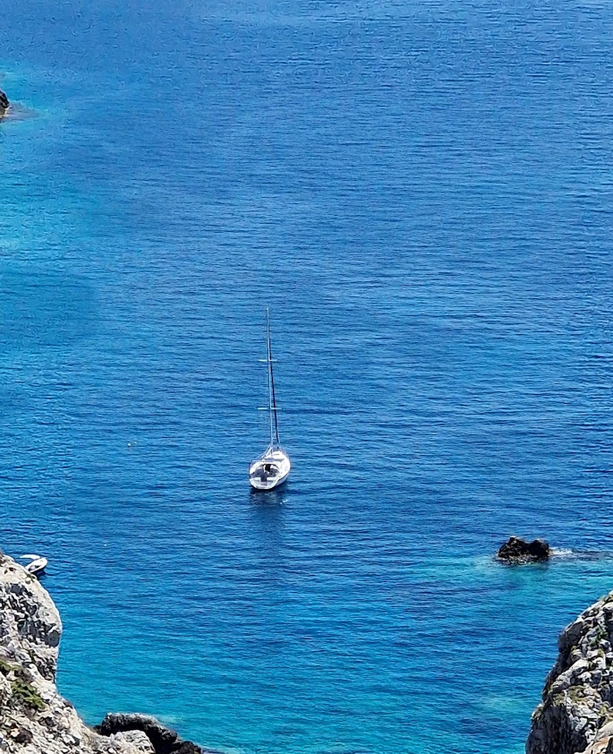 A clear view of a sailboat on calm blue waters with rocky cliffs in the foreground.