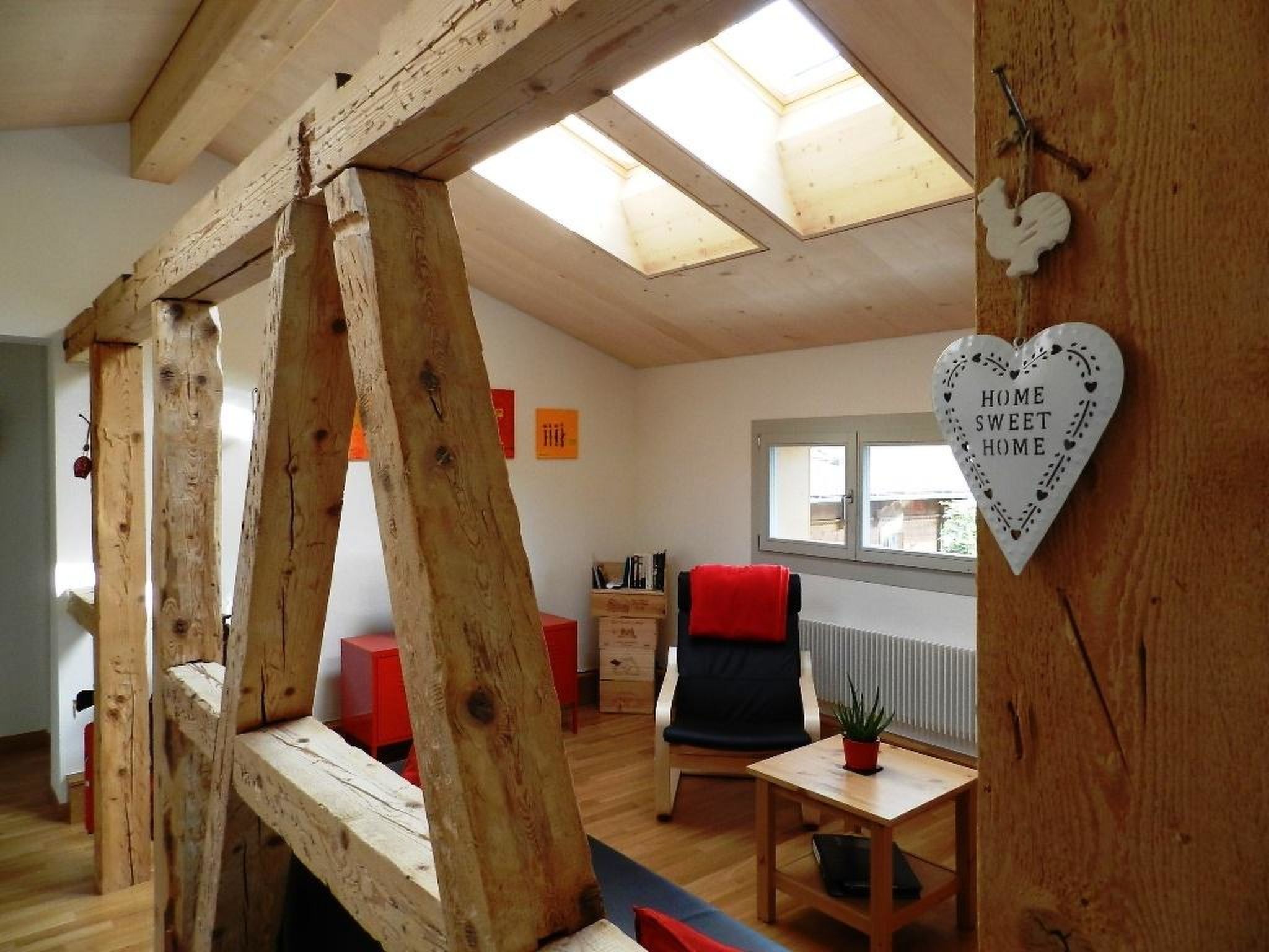 Une pièce avec une structure en poutres apparentes, une chaise rouge, une petite table avec une plante et un décor en forme de cœur avec un texte.