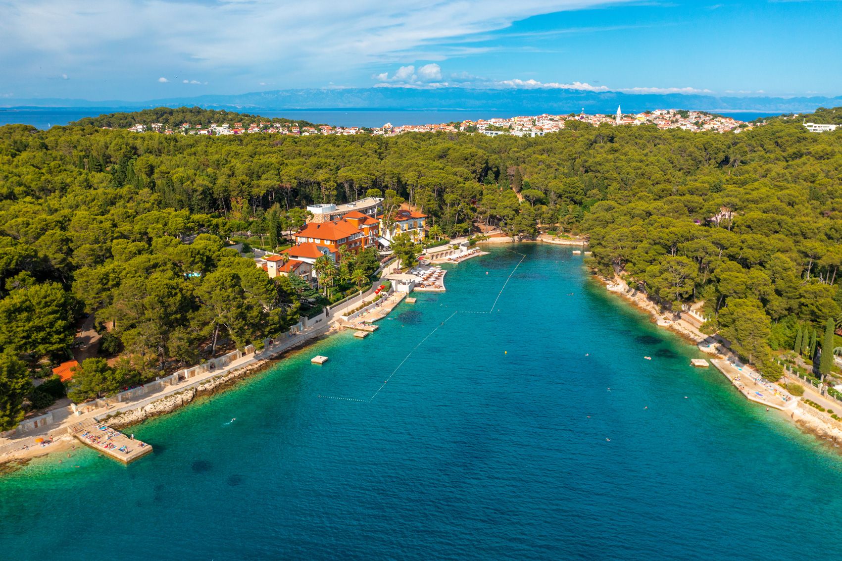 An aerial view of a coastal town with a marina, surrounded by dense green forests and a clear blue sea.