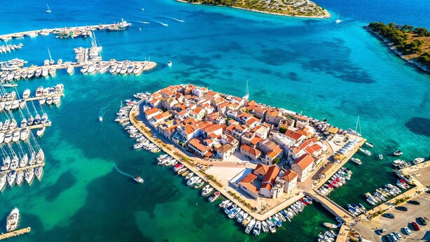 Vista aérea de un pueblo costero con una plaza central, barcos amarrados en el puerto deportivo y aguas azules cristalinas.