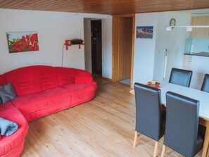 A living room with a red sofa, a dining table with chairs, and a kitchen area in the background. - Burkhalter Appartement/Fewo, Dusche oder Bad, WC 4-Bettwohnung by Interhome (Zweisimmen)