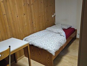 A small, single-bed room with a wooden headboard, a bedside table, and a red bedspread. - Burkhalter Appartement/Fewo, Dusche oder Bad, WC 4-Bettwohnung by Interhome (Zweisimmen)