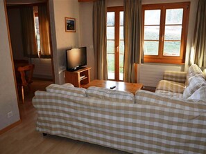 A living room with a checkered sofa, a television on a wooden stand, a dining area with a chair, and a window with curtains. - Jacqueline 1 Appartement/Fewo, Dusche oder Bad, WC 6-Bettwohnung by Interhome (Gstaad)