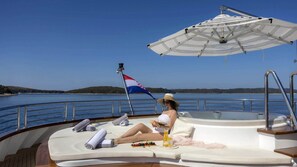 Eine Person entspannt auf einem Bootsdeck mit einer Flagge, einem Sonnenschirm und einem Getränk.