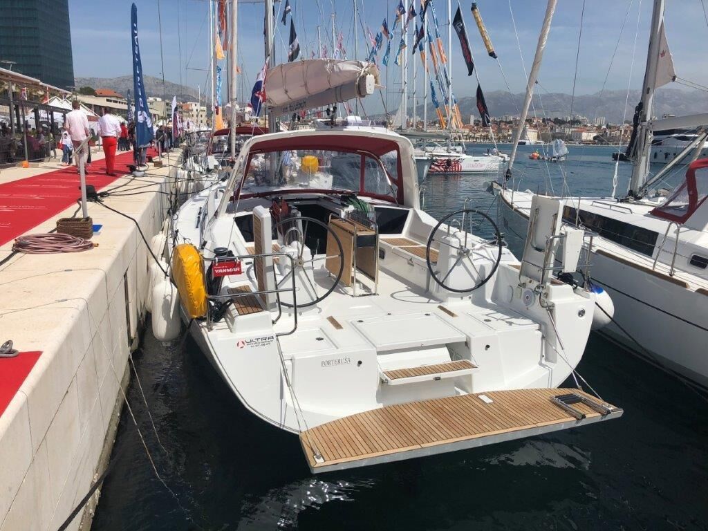 Ein Boot liegt in einer Marina festgemacht, mit rotem Teppich, einem Holzboden und einer Stadtkulisse im Hintergrund.