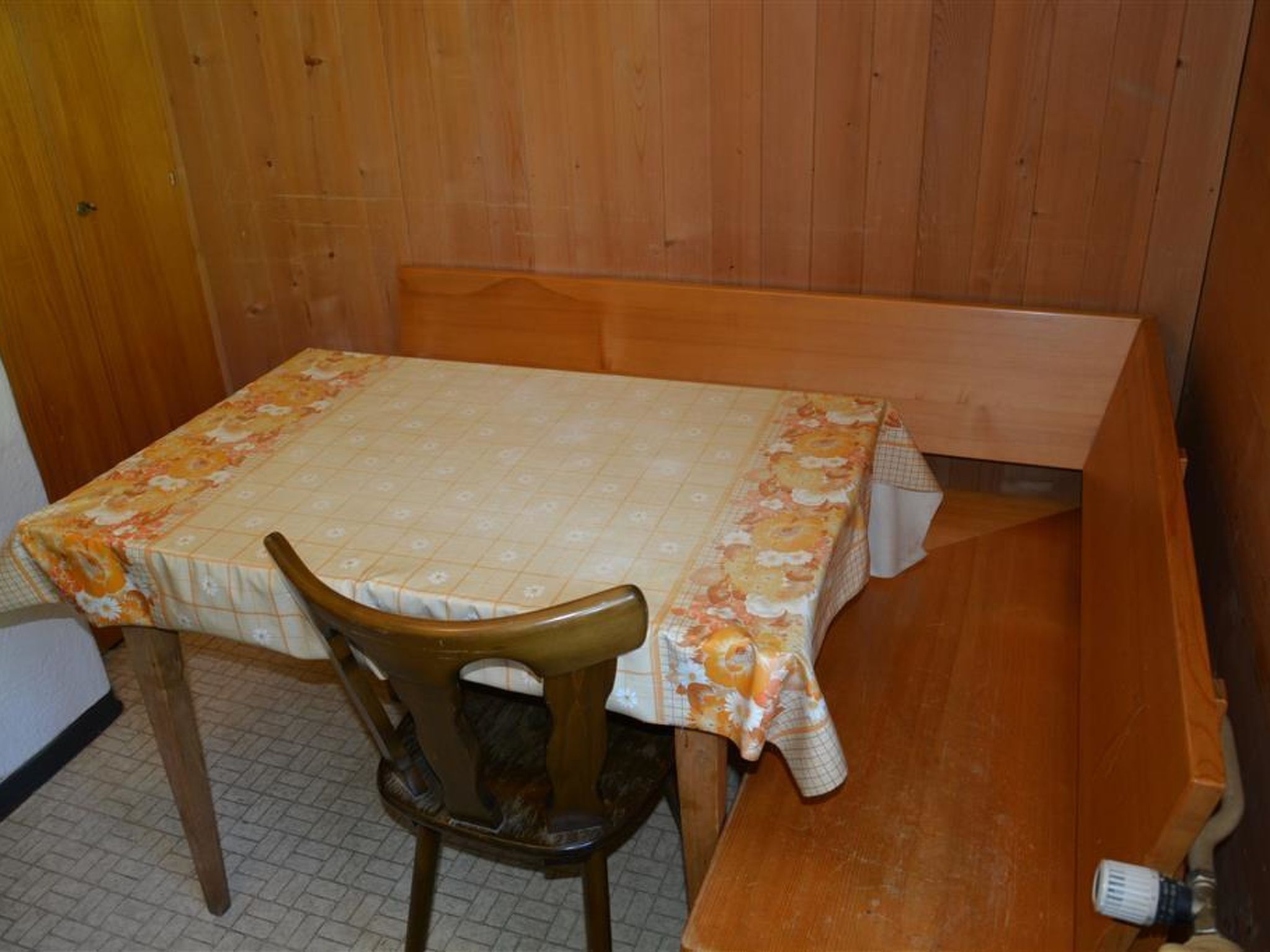 Uma pequena mesa de madeira com toalha de mesa floral, um banco de madeira e uma cadeira em um quarto com paredes de madeira.