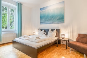 A hotel room with a bed, a nightstand with a lamp, a chair, and a large window with curtains. - Stadt Land Fluss Apartment 2 (Regensburg)