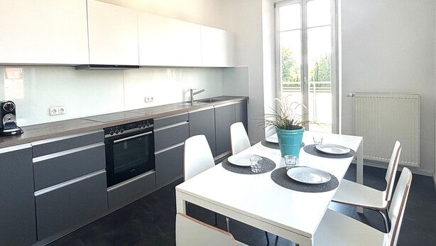 A modern kitchen with white cabinets, a built-in oven, and a dining area with a white table and chairs. - Stadt Land Fluss Apartment 2 (Regensburg)